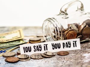 Image of money next to a tipped over swearing jar. Sign says "You say it you pay it"