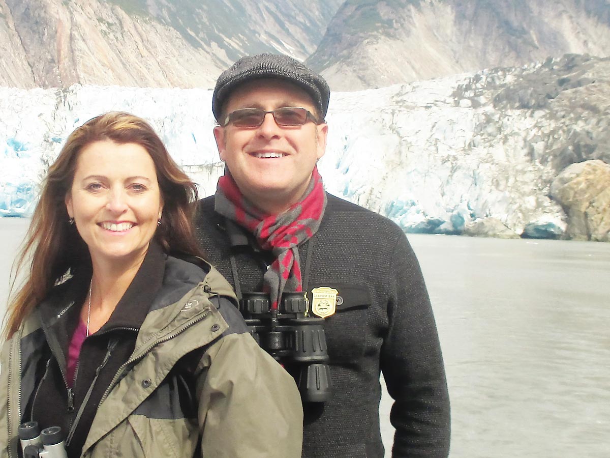 Laura Sable and Bill Wiemuth standing on a boat in Alaska. They offer virtual cruises.
