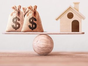 photograph of money sacks balanced on a plank balanced on a ball, with a house on the other end of the plank