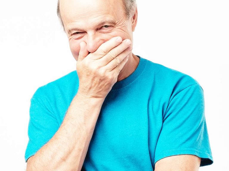 Senior man in a blue t-shirt smirking while covering his mouth with his hand.